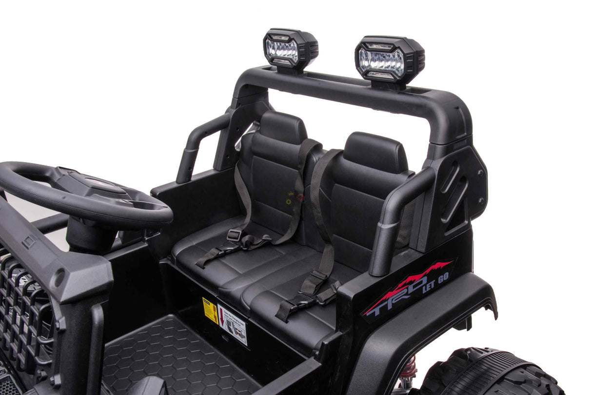 A close-up of the Black 24V/4WD Xtreme Ride on Truck shows its interior featuring two black leather seats with seatbelts, a steering wheel, and a roll bar. Mounted lights on top and Eva Wheels complete the rugged, adventurous look.