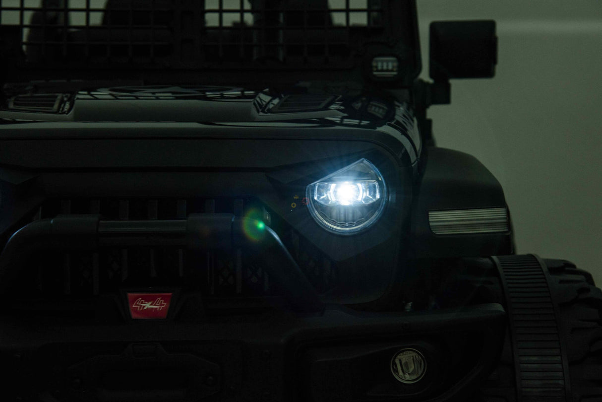 Close-up of the Black 24V/4WD Xtreme Ride on Truck’s front, emphasizing a bright headlight. The grille is visible with a red badge underneath, highlighting its rugged design and advanced Eva Wheels in dim lighting.