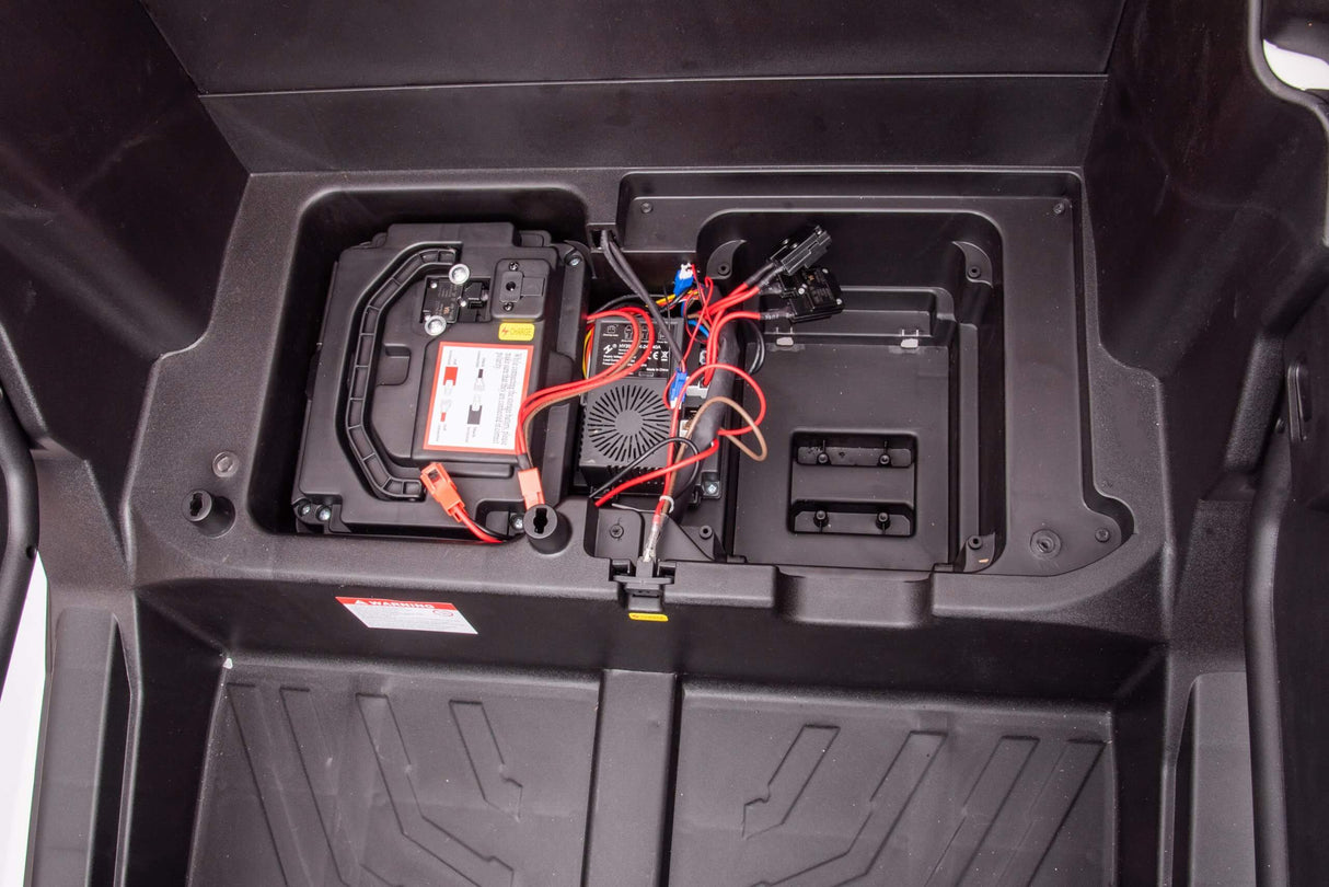 In the open compartment of a Rose 2x24V/4WD Official Can-Am Maverick Ride on Buggy, LX Performance, a car battery is connected to wires on the left. To the right is space with smaller components. The black plastic finish features geometric patterning at the bottom.