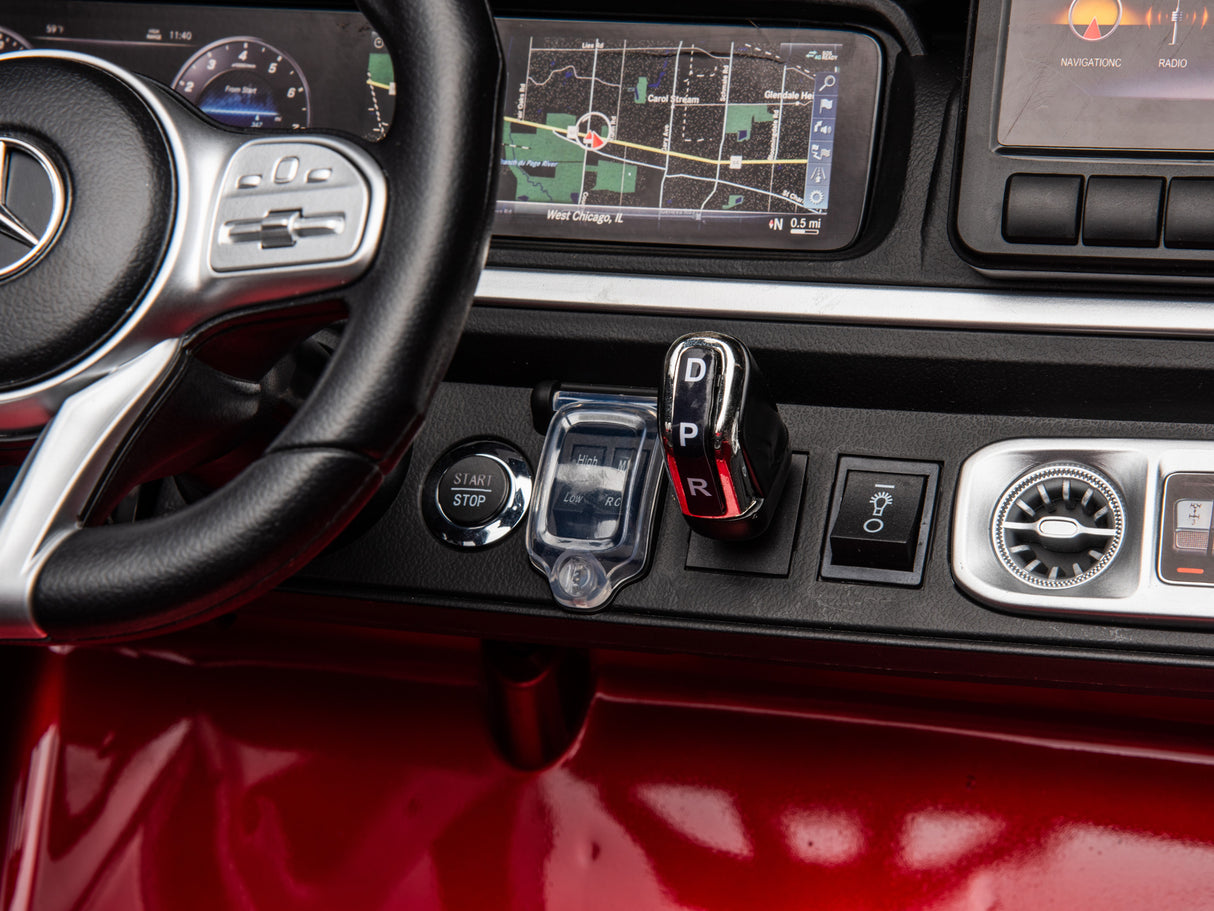 Close-up of the Red Metallic 2x24V/4WD Platinum XXL Mercedes G63 Ride On Truck dashboard, showcasing a steering wheel, gear shift with P, R, N, D, and 1, start/stop button, climate controls, navigation screen, and sleek black surface with red trim.