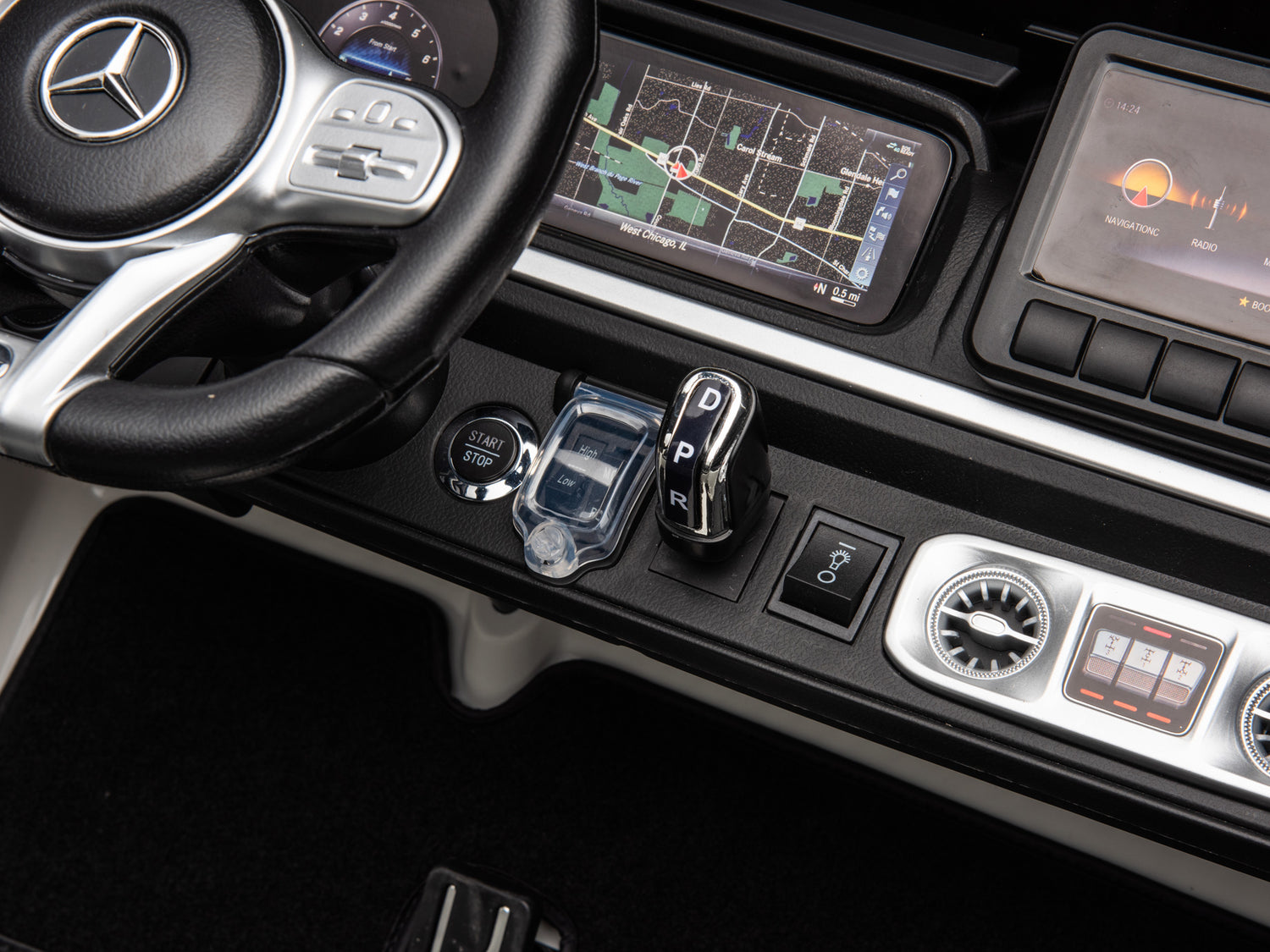 Close-up of a White 2x24V/4WD Platinum XXL Mercedes G63 Ride-On Truck dashboard featuring a logo-emblazoned steering wheel, infotainment screen with map, start/stop button, gear shift, and audio-climate control panel. Enjoy remote control access to this 4-wheel drive adventure.