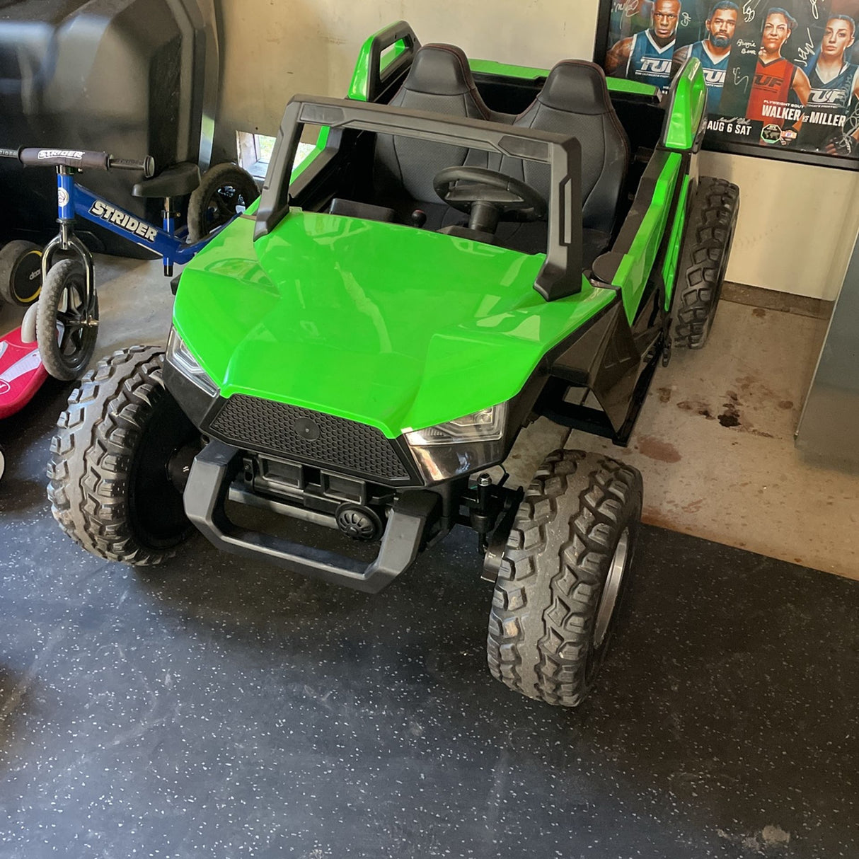 A bright green 24V/4X4 Dune Buggy Ride On UTV with large black rubber wheels is parked on a garage floor. Nearby sits a blue balance bike and a red bicycle. Posters adorn the walls in the background, enhancing the fun atmosphere.