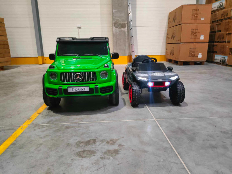 A Green Mercedes G63 XXL 24V Kids Ride-On Truck – Platinum Edition with MP4 features dual batteries, 4WD, EVA tires, and leather seats parked next to a black toy Audi in an industrial storage space filled with boxes and a ladder, offering remote control luxury car vibes in miniature.