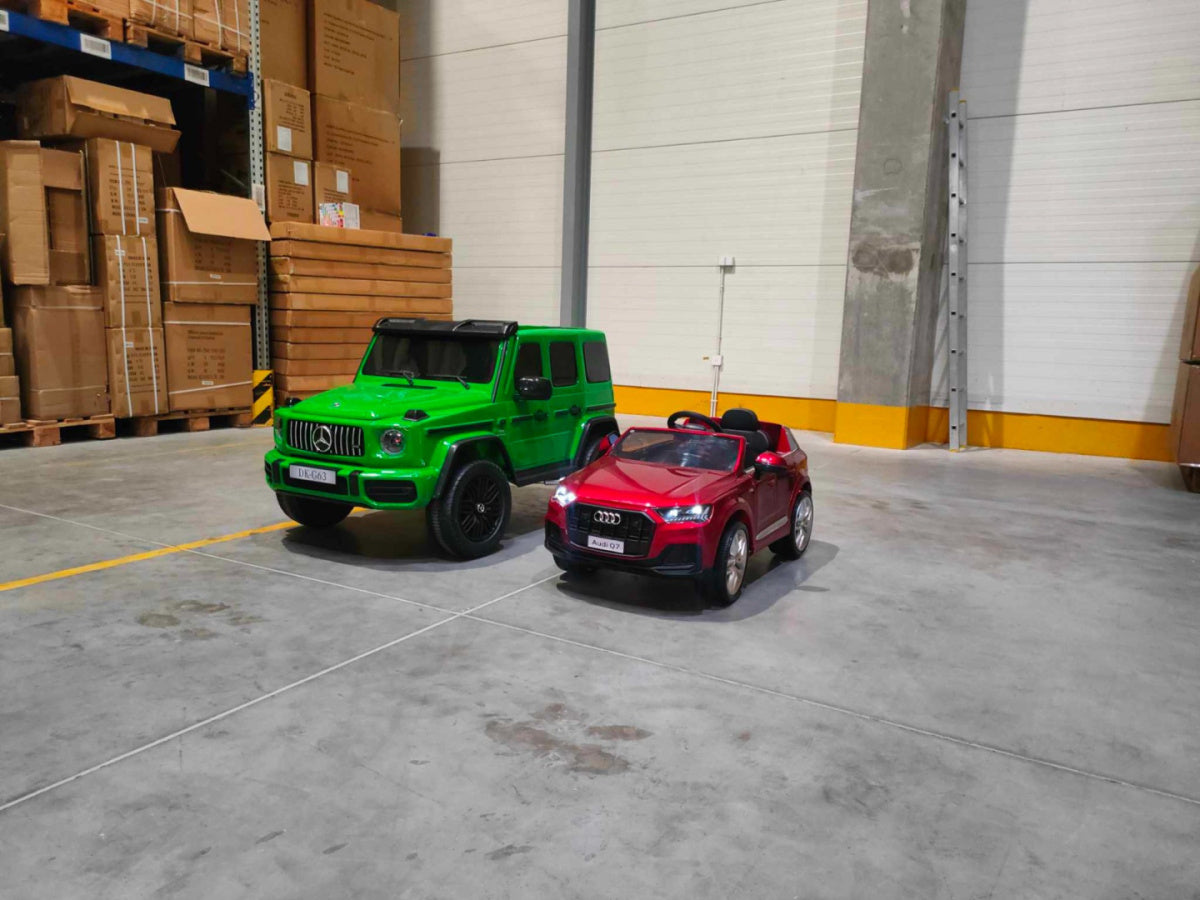 A green Mercedes G63 XXL 24V Kids Ride-On Truck with MP4, dual batteries, 4WD, and a red Audi-style convertible are parked indoors on concrete. Cardboard boxes are stacked on wooden pallets in the background.