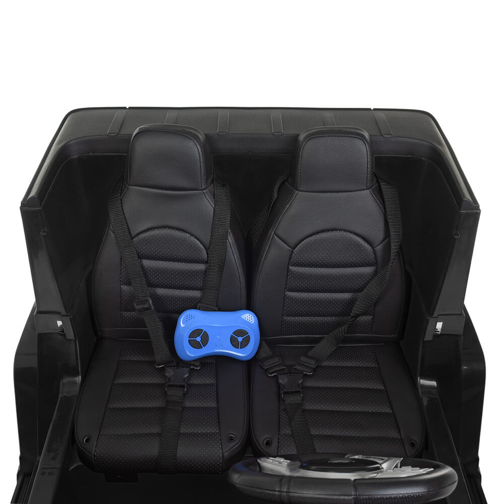 The luxurious interior of the Official Mercedes Benz G63 Kids Ride On Car features two black leather seats with seat belts. A blue remote control hints at its advanced 24V battery-powered system, and the textured pattern adds elegance as a steering wheel peeks into view.