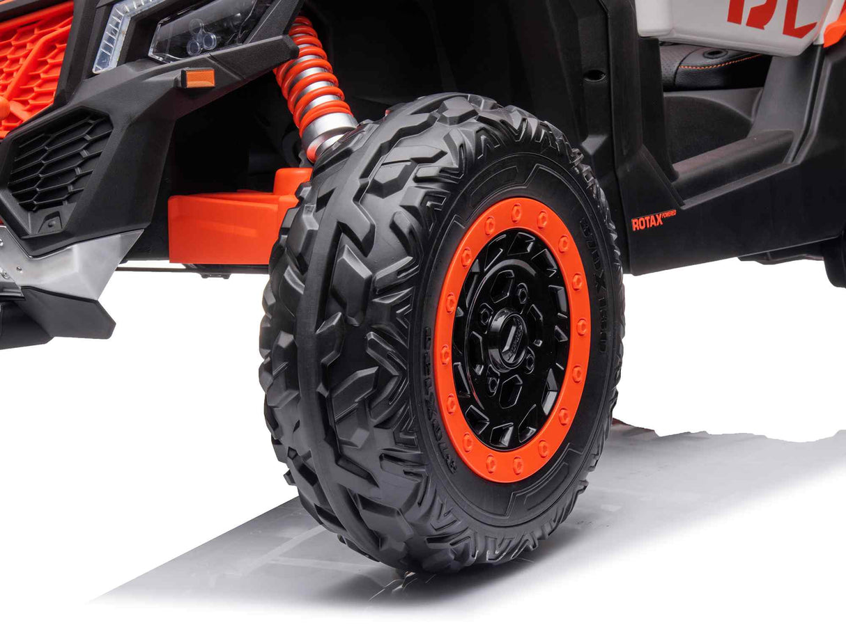 Close-up of a rugged, deeply treaded front tire on the 2 Seater Official 2x24v Can-Am Maverick RS Ride on Buggy, LX Performance 4WD Edition. The orange and black rim and matching orange suspension springs stand out against the white background, highlighting its impressive design.