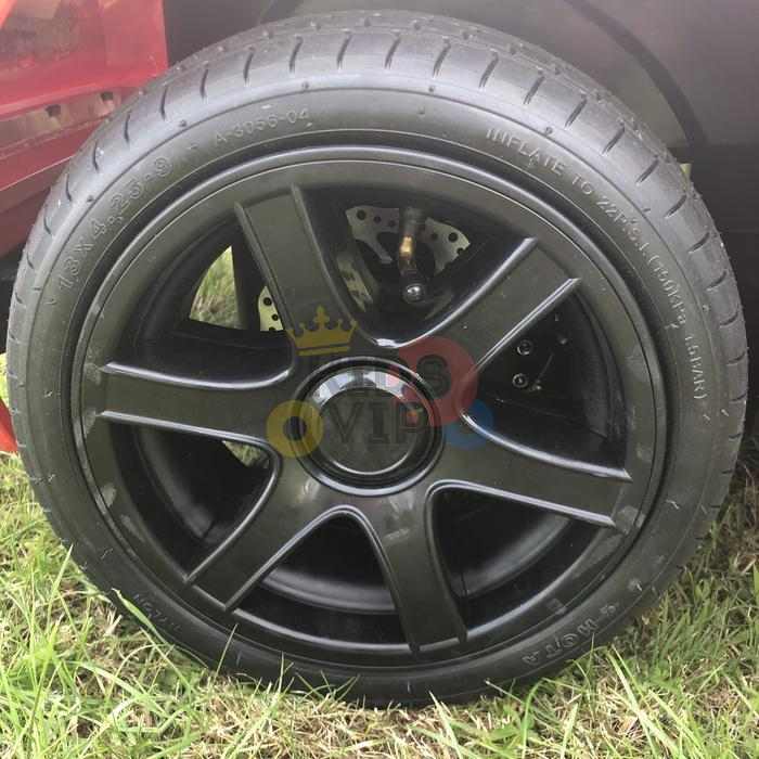 Close-up of a black car wheel with five spokes and visible tread, designed for the Orange 24V XXL SuperSport Big Kids Ride On Car. The wheel rests on grass, featuring an OVH logo encircled by a yellow ring and crown design.