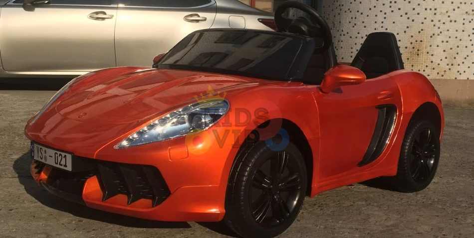 An Orange 24V XXL SuperSport Big Kids Ride On Car with a 180W brushless motor and real tires is parked stylishly on concrete with black accents and side mirrors, while a larger silver car appears partly visible near a building in the background.