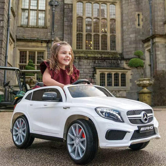 A young girl with a big smile sits in a toy white Mercedes-Benz car. She is outdoors in front of a large, historic building with tall windows and stone walls. The car is styled like an AMG model.