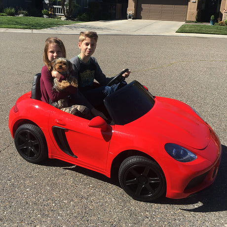 A boy drives and a girl cuddles a small brown fluffy dog as they sit in a charming red 24v/180w XXL Super Ride styled like a convertible sports car. The vehicles impressive rear axle and motor assembly quickly propel them past houses and driveways.