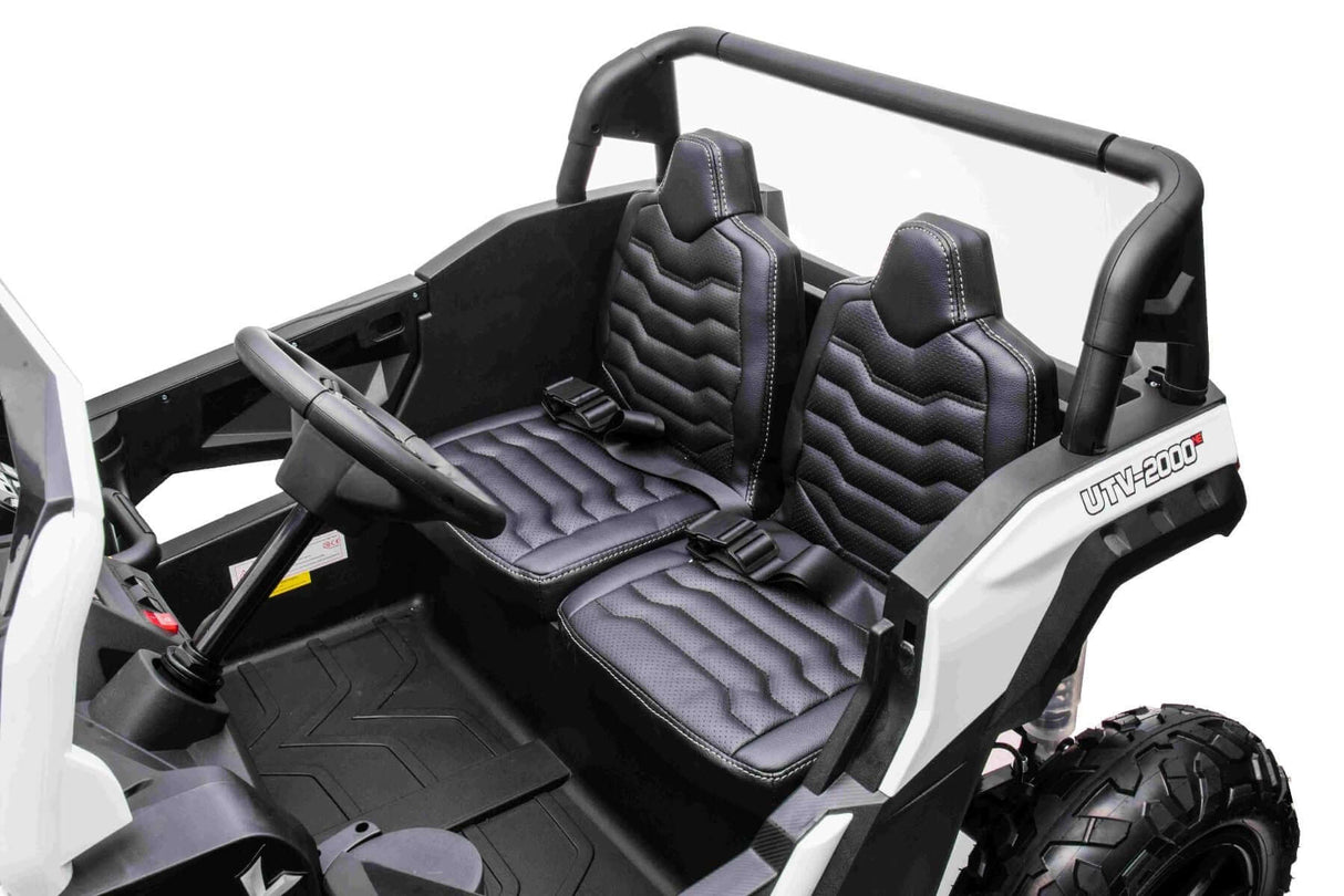 The image showcases the interior of a White 24V/180W XXL Big Kids Blade XR Ride on Buggy. It features two black cushioned seats with stitching, a steering wheel, and a minimalist dashboard. The white exterior has striking black accents and inflatable rubber tires, enhancing its off-road appeal.