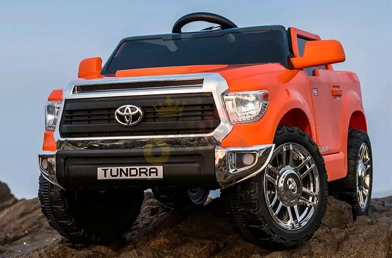 An Orange 12V Licensed Upgraded Toyota Tundra Kids Ride On Truck is parked on rocky terrain, featuring detailed styling, a large black grille with chrome accents, and shiny wheels, closely resembling a rugged full-sized pickup.