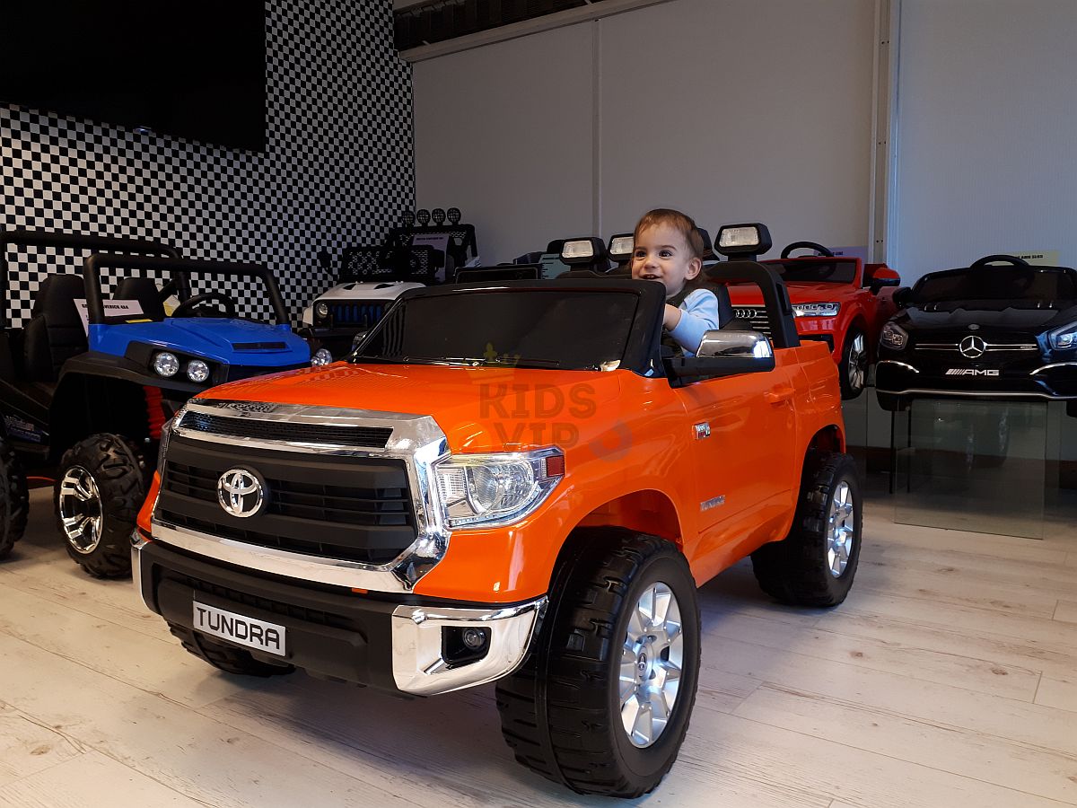 A child eagerly grips the steering wheel of an orange 12V Licensed Upgraded Toyota Tundra 1 Seater Kids Ride On Truck with RC. In the background, a showroom displays other toy cars against a checkered wall and wooden floor, promising thrilling adventures for young drivers.