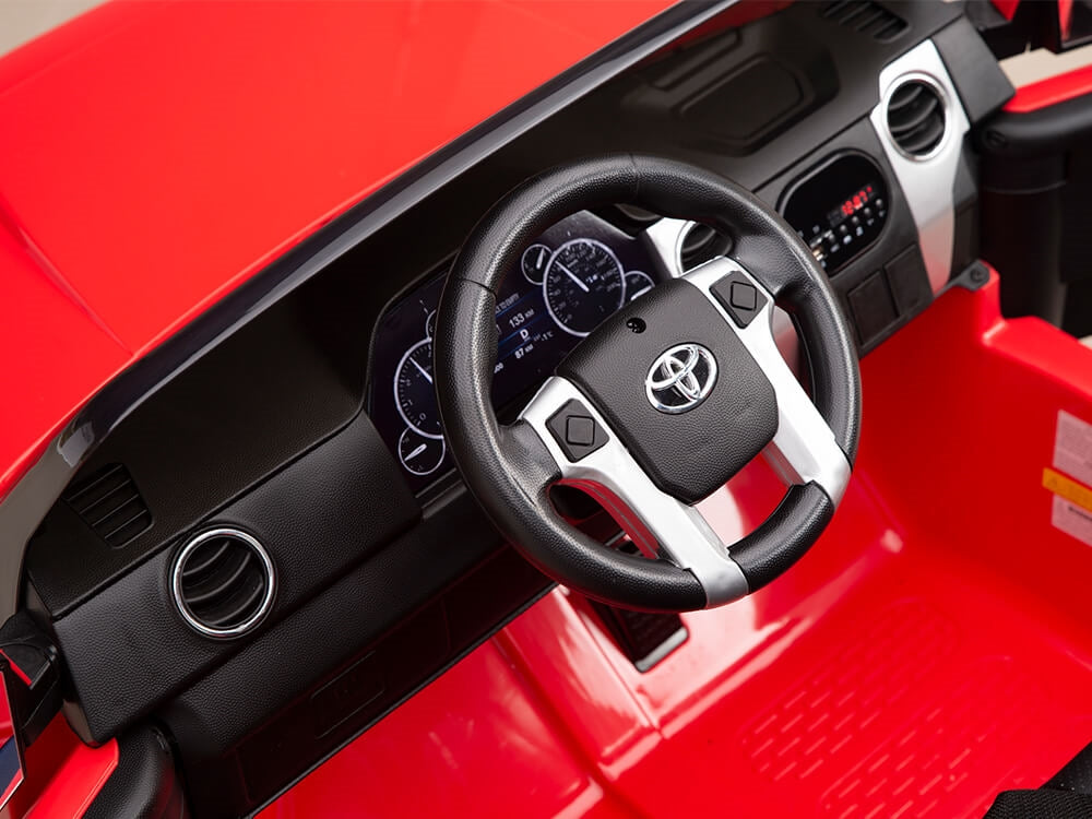 A close-up of the Red 12v Licensed Toyota Tundras dashboard shows a black and silver steering wheel with a Toyota logo. The digital display and air vents are seamlessly integrated into the red interior of this 2-seater Kids Ride On Truck.