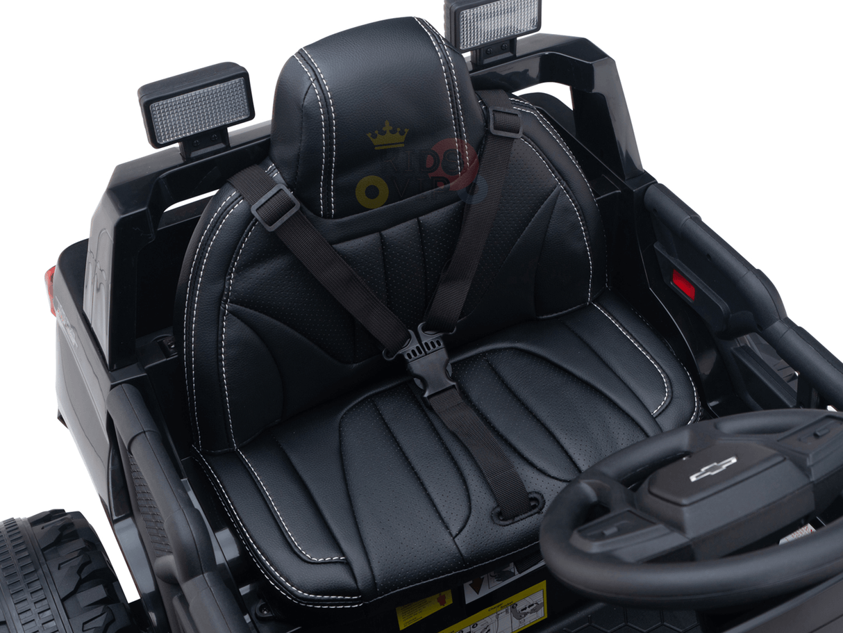 The Black Official 24V Chevrolet Silverado Lifted Kids Ride-on Truck features a black leather seat with white stitching, a logo on the small steering wheel, and a seatbelt. Its cushioned and detailed design offers comfort for young drivers.
