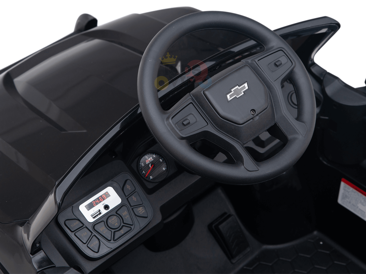 Close-up of a black toy cars interior, resembling the Black Official 24V Chevrolet Silverado Lifted Kids Ride-on Truck. It features an iconic logo on the steering wheel and a dashboard with a speedometer and control buttons on a textured surface.