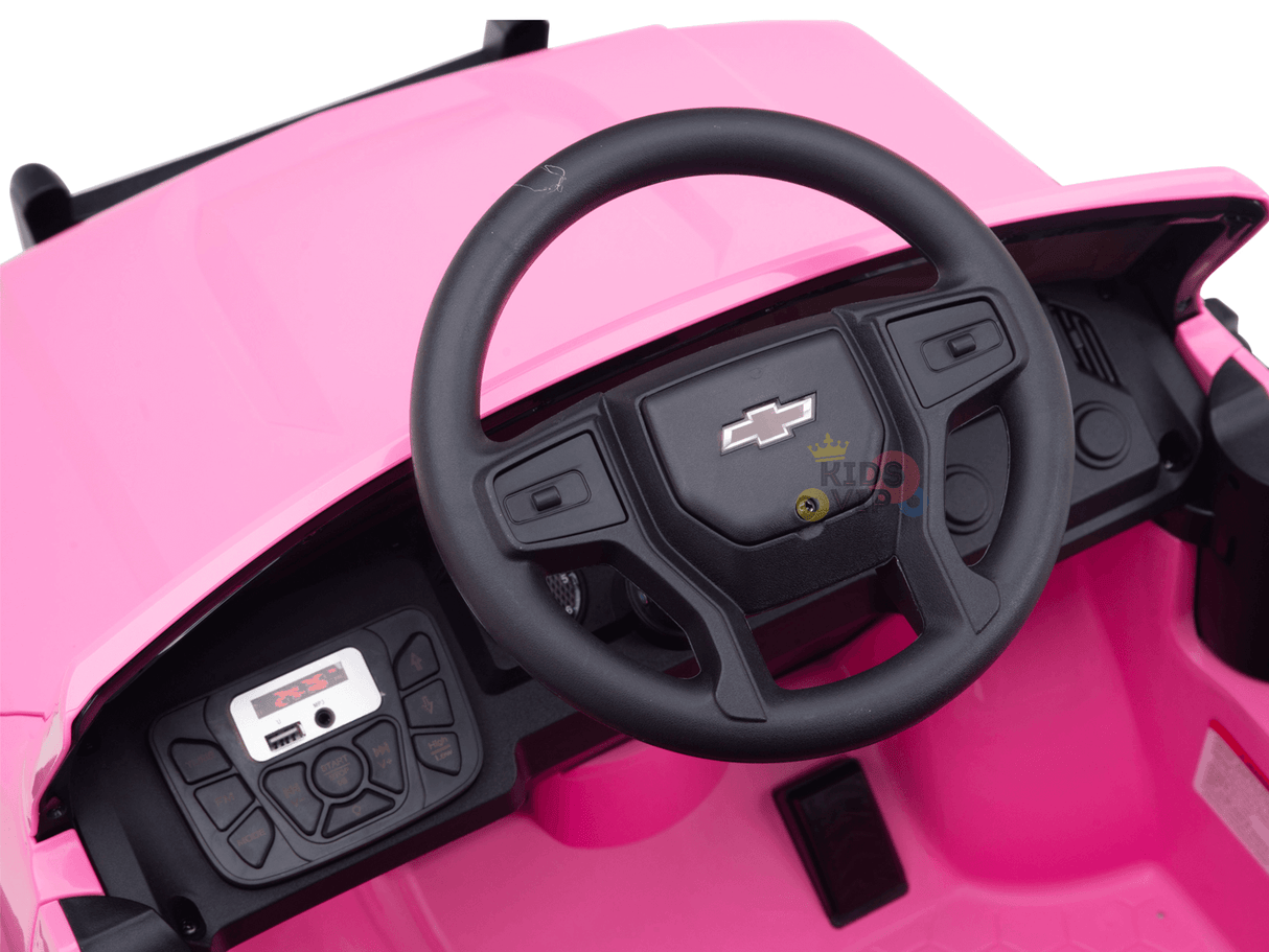 The image showcases the interior of a Pink 24V Chevrolet Silverado Kids Licensed 1 Seater Ride On Lifted Truck. It features a black steering wheel with a Chevrolet logo, mini dashboard with buttons, and digital display on the left, resembling an authentic Chevrolet Silverado.