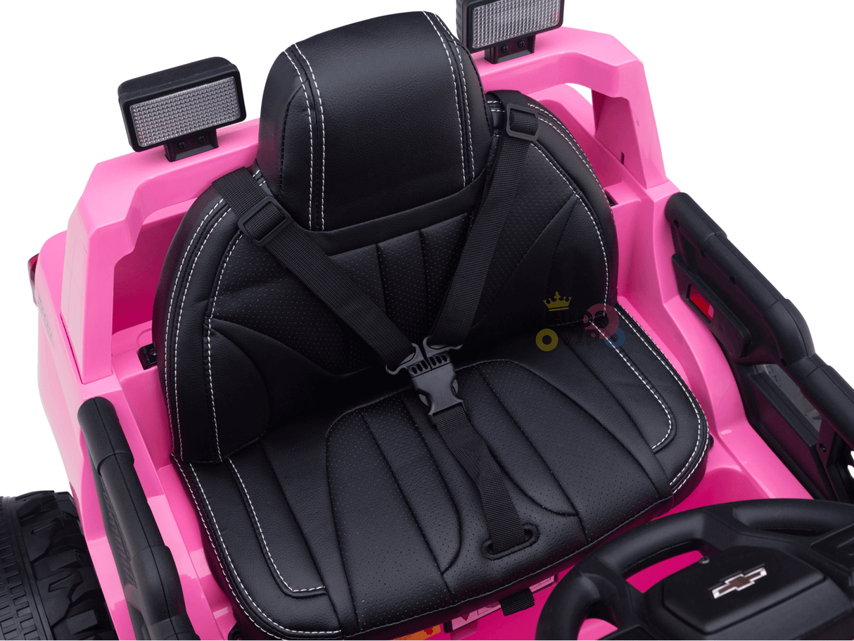 The Pink 24V Chevrolet Silverado Kids Licensed 1 Seater Ride On Lifted Truck features a charming interior with a black seat, white stitching, seatbelt, small steering wheel, and top light fixtures. Its streamlined dashboard ensures ease of use for young drivers.