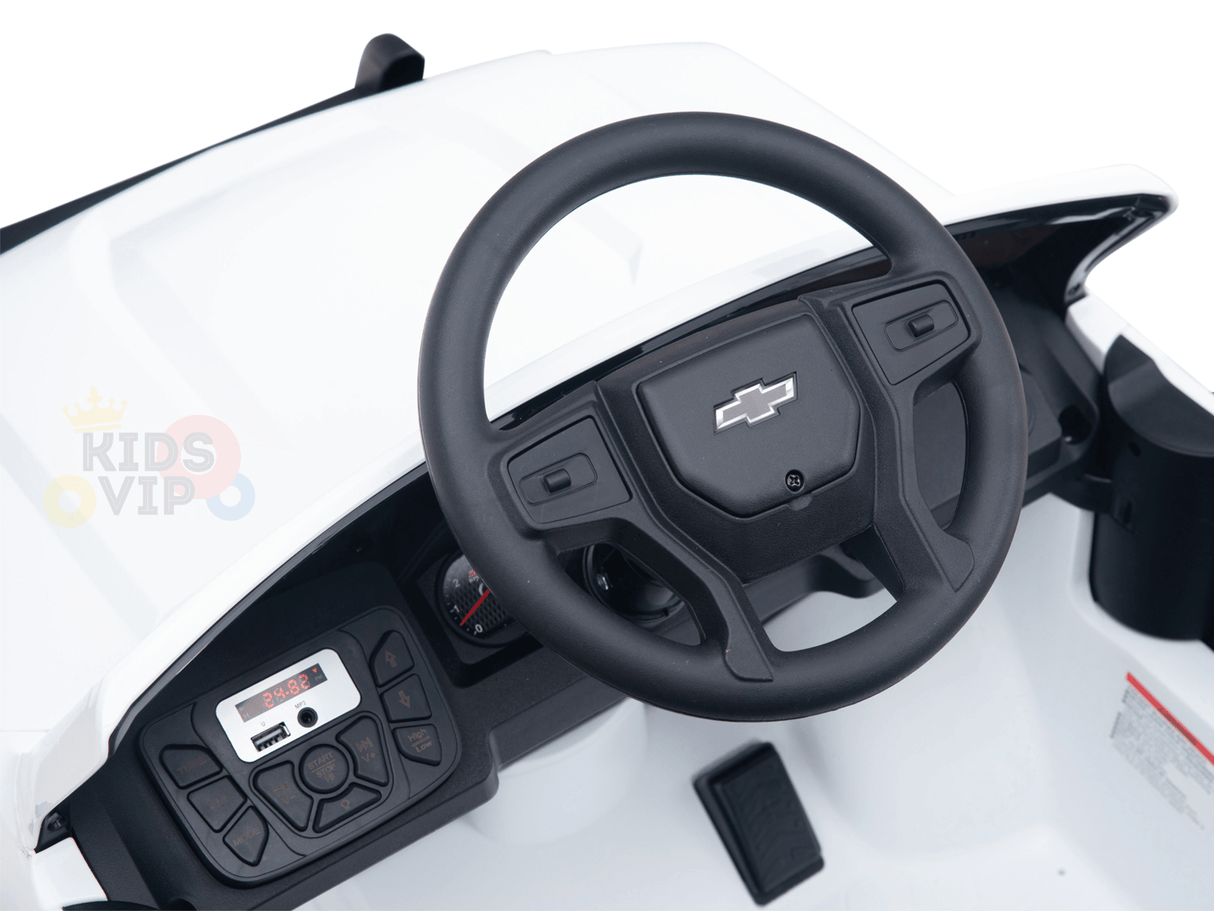 A close-up of the White 24V Chevrolet Silverado Kids Licensed 1 Seater Ride On Lifted Truck dashboard reveals a black Chevrolet-logo steering wheel, intricate control panel, and digital display. The interior features realistic detailing, with a primarily white design accented in black.