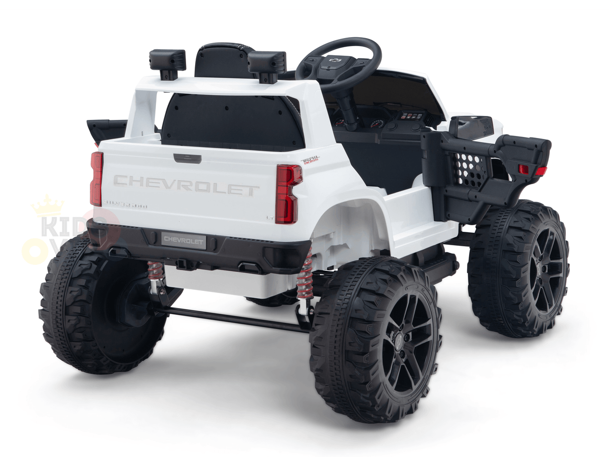 The rear angle of the White 24V Chevrolet Silverado Kids Licensed 1 Seater Ride On Lifted Truck with Remote shows oversized black tires, a detailed dashboard, and realistic elements like a tailgate and side mirrors. The toy is set against a white background.