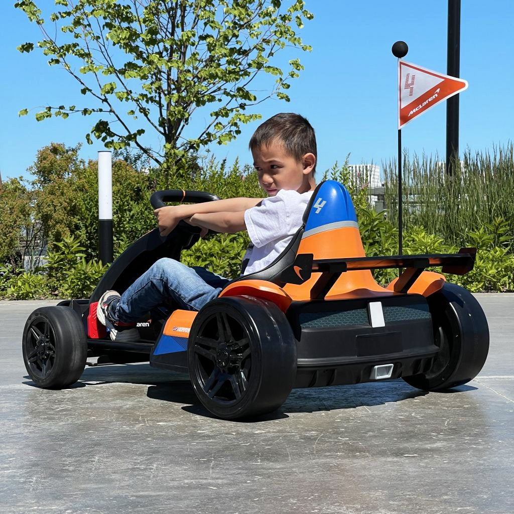 A child in a blue shirt and jeans speeds an Orange 24V Official Complete Edition McLaren Super Drifting Go-Kart down a paved path, featuring a rear spoiler, black wheels, and a flag with the number 4. Trees and grass blur in the background as the young racer zooms past.