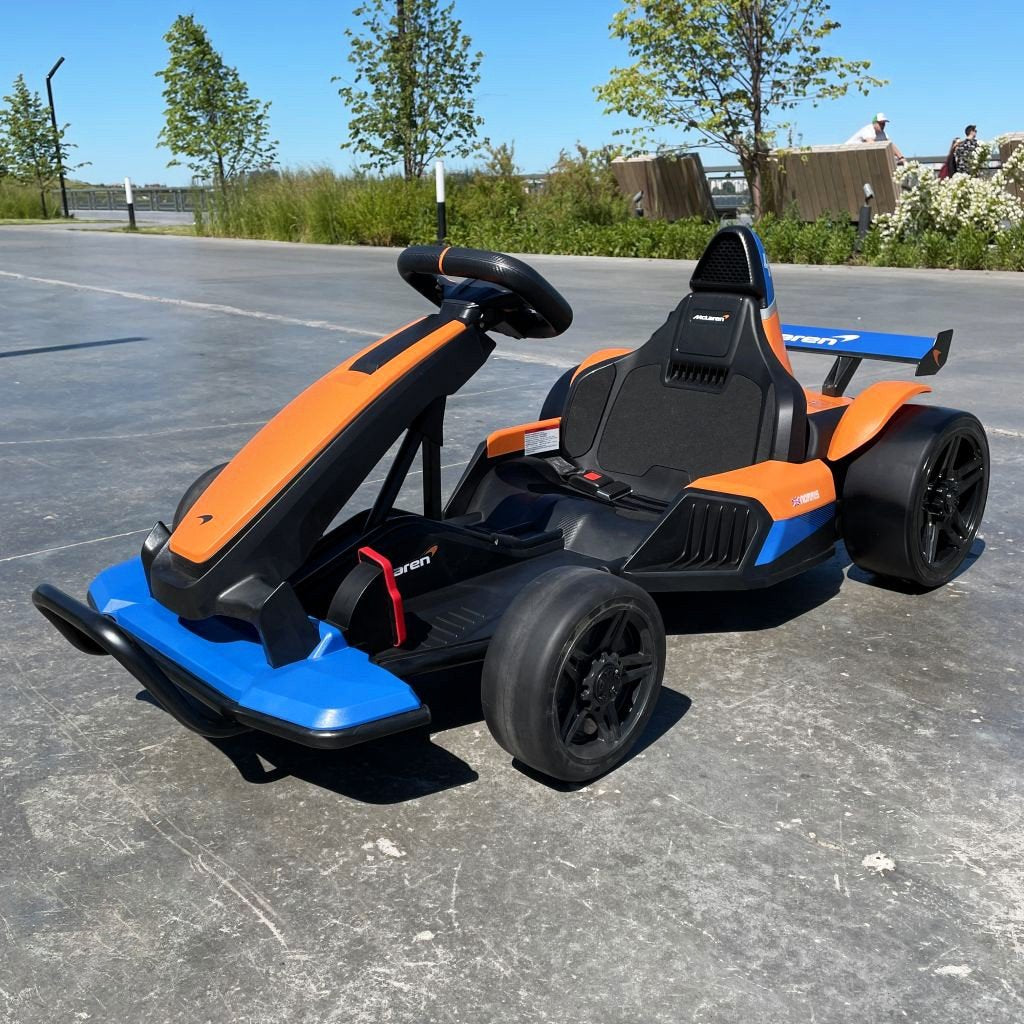 An Orange 24V Official Edition McLaren Super Drifting Go-Kart with a futuristic design and black wheels is parked on concrete, accentuated by orange and blue colors. It features a low seat for optimized drifting, set against trees and grass under a clear blue sky.