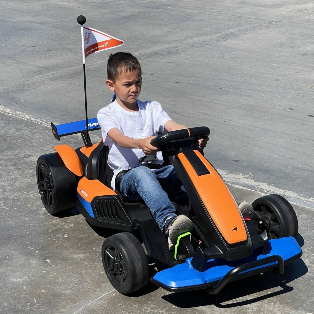 A young boy in a white shirt and jeans drives an Orange 24V Official Complete Edition McLaren Super Drifting Go-Kart on concrete, featuring a small flag. He looks focused as he steers.