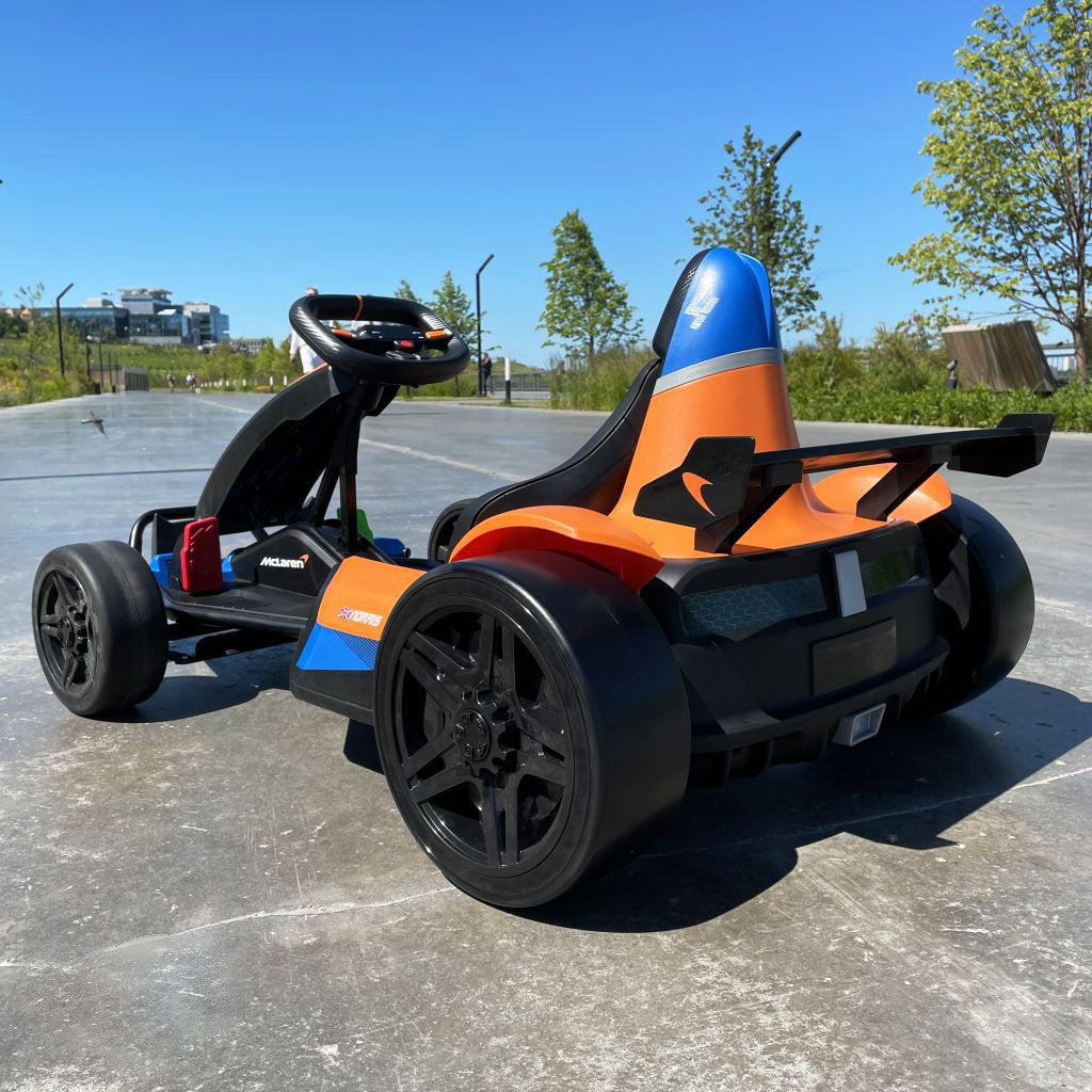 Close-up of an Orange 24V Official Complete Edition McLaren Super Drifting Go-Kart with an orange and blue design, parked on concrete. The kids ride-on has a steering wheel, seat, and large black wheels. Trees and buildings can be seen in the background under a clear blue sky.