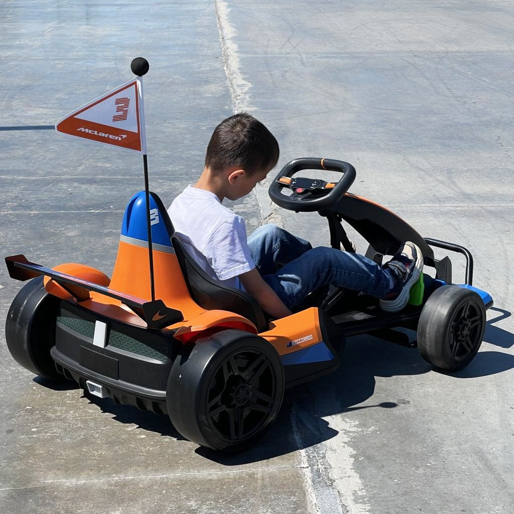 A child in a white shirt sits excitedly in the Orange 24V Official Complete Edition McLaren Super Drifting Go-Kart, its small flag fluttering in the sun. This go-kart, ideal for kids ride-on adventures, is ready for drifting thrills on a paved surface.