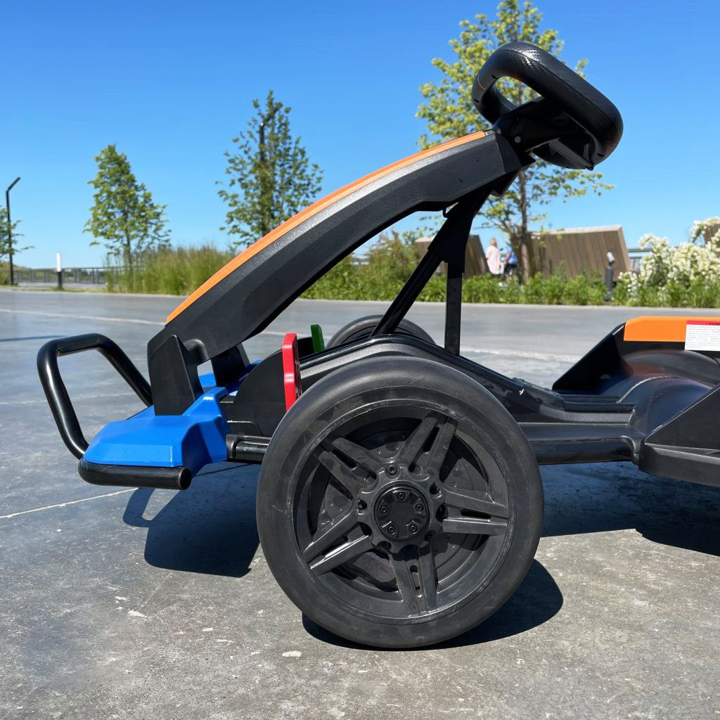 Close-up of an orange 24V Official Complete Edition McLaren Super Drifting Go-Kart with large wheels on a concrete surface. The background shows a clear blue sky, green trees, and a fence, suggesting a park setting ideal for this exciting kids ride-on experience.