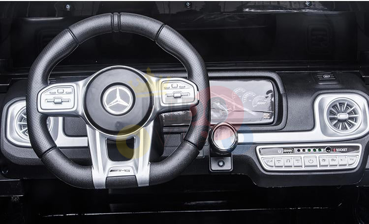 A close-up of the dashboard in the Mercedes Benz G63 kids ride-on car showcases the luxury logo on the steering wheel. Featuring control buttons and air vents, it offers a modern, predominantly black design with metallic accents, mirroring high-end automotive style.