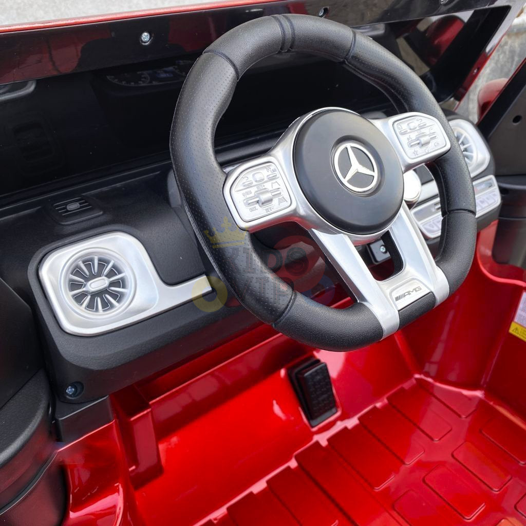 A close-up of this 2-seater 4x4 Official Mercedes-Benz G63 ride-on cars steering wheel and dashboard showcases a Mercedes logo. The sleek black and metallic interior details mirror the real G63, with controls, vents, and a shiny red plastic floor, all powered by a robust 24V battery.