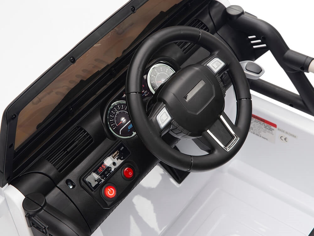 Interior of the White 12V Sport Edition Trailcat Big Wheels Kids Ride on Truck features a black steering wheel, dashboard with gauges, two buttons including a red power button, simple control panel, and tan-colored windshield for visibility.