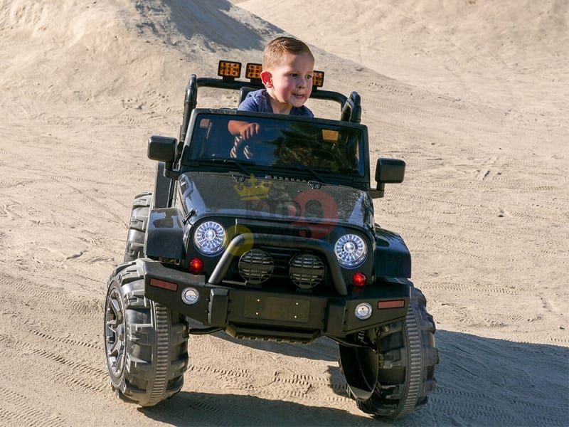 A young child joyfully drives a blue 12V Sport Edition Trailcat Big Wheels Kids Ride on Truck with RC across the sandy terrain. Smiling, the child grips the wheel as large tires conquer uneven ground, trailing sand and a small hill in the background.