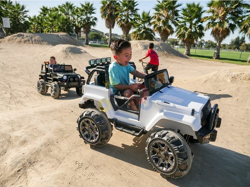 Two children navigate their Blue 12V Sport Edition Trailcat ride-on trucks on a dirt track with ramps. A boy in a white vehicle leads as the one in black follows closely. Meanwhile, a third child pedals a bicycle in the background, surrounded by palm trees and lush greenery.