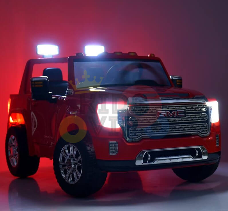 The Red 2x12V Licensed 4WD GMC Sierra Kids Ride On Car with RC is displayed in a dimly lit setting, featuring illuminated headlights and roof lights. Positioned at an angle, it showcases its detailed grille and shiny wheels.