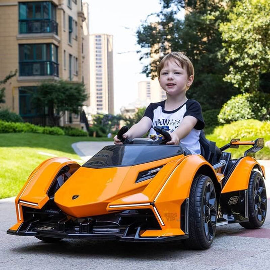 A young child wearing a white and black shirt smiles while driving an orange toy sports car on a path. The background features tall residential buildings and green trees.