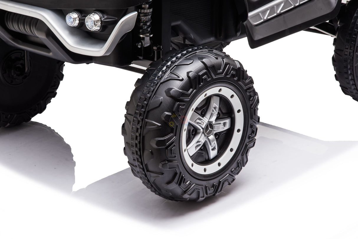 A close-up of the White 12V/4X4 Official Mercedes Unimog Junior Ride On UTV reveals a rugged tire with deep treads, reminiscent of an off-road vehicle. Its metallic rim features a five-spoke design, and the kids ride-on boasts visible suspension and a sleek black exterior with a polished chrome bumper.