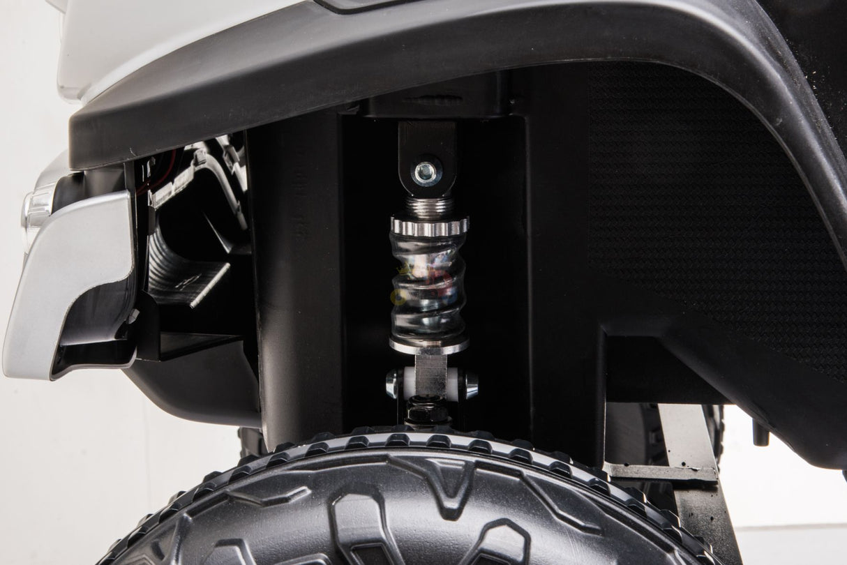 Close-up view of the suspension system on the Black Official 12V Mercedes Unimog Junior 4X4 Kids Ride-On UTV, showing a coil spring and shock absorber above an off-road tire, emphasizing its durable design and powered by a 12V battery.