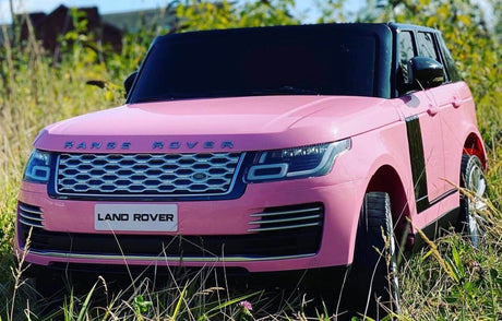 A pink 12V Range Rover HSE toy car, featuring detailed headlights and a Land Rover plate, sits on the grass surrounded by tall plants. This ride-on car is perfect for adventurous kids seeking a thrilling journey with the aid of a superior remote control.