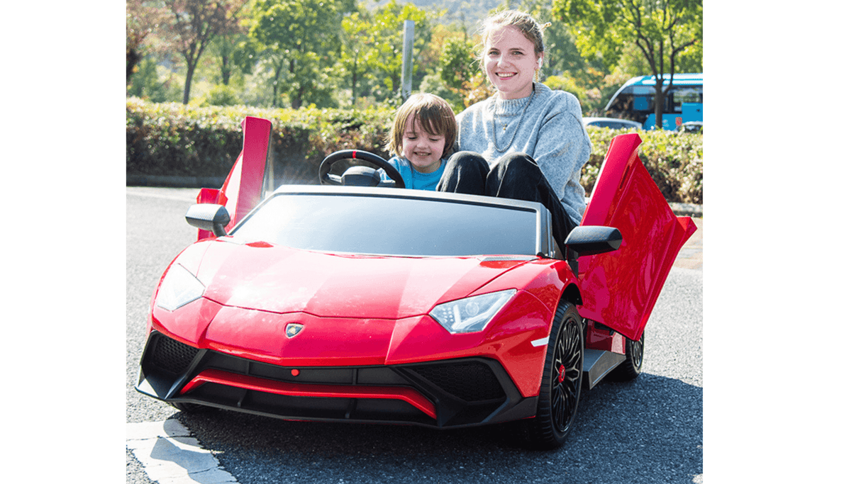 A woman and child enjoy a sunny day in their Red 24V/180W Lamborghini SV Limited XXL Edition ride-on toy, its doors wide open. The child playfully holds the steering wheel while lush trees and a blurred vehicle frame this vibrant Kids VIP moment with speeds up to 10MPH!.