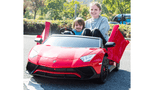 A woman and child enjoy a sunny day in their Red 24V/180W Lamborghini SV Limited XXL Edition ride-on toy, its doors wide open. The child playfully holds the steering wheel while lush trees and a blurred vehicle frame this vibrant Kids VIP moment with speeds up to 10MPH!.