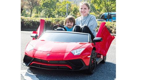 A smiling woman and child enjoy a sunny day in their Red Giant Official Limited Edition Lamborghini SV 24V/180W, doors open, surrounded by greenery. The ride-on supercar hits up to 10MPH!!!, creating an XXL edition of Kids VIP fun with a parked car as the backdrop.