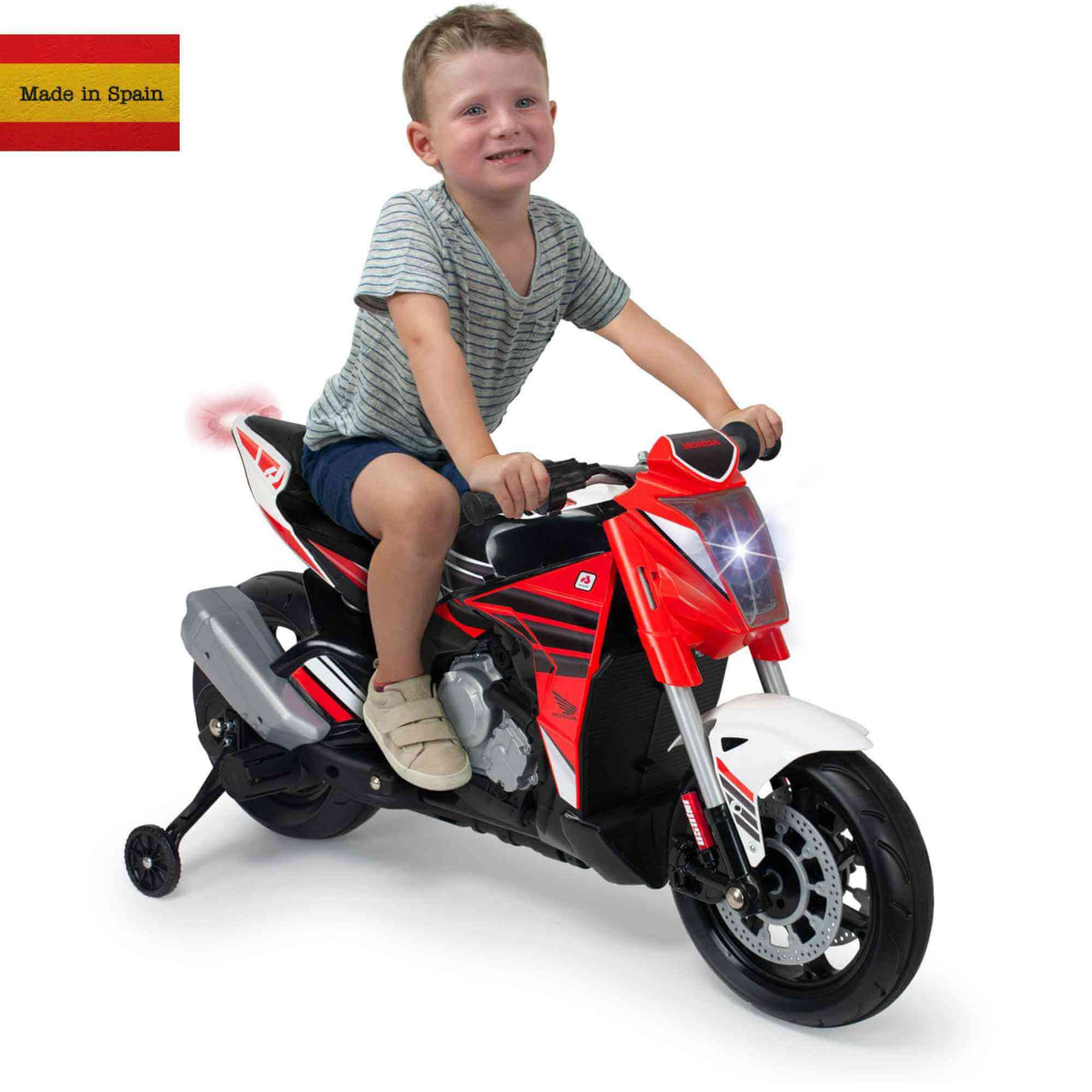 A child rides a Red INJUSA 12V Honda Naked Edition ride-on bike with training wheels in a gray striped shirt and shorts. The motorcycle has a headlight, black wheels, and displays a small Spanish flag with Made in Spain in the top left corner.