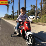 A young boy rides a small red bike with training wheels along a sunny road lined with palm trees, wearing a white and gray striped shirt. He looks focused as if imagining its a Red INJUSA 12V Honda Naked Edition Ride On Bike/Motorcycle for Kids—proudly Made in Spain.