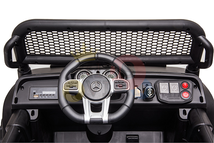 The image displays the interior of a Camo 12V/4X4 Official Mercedes Unimog Junior Ride On UTV, highlighting a detailed steering wheel with logo, dashboard controls, and a front mesh barrier. The dashboard is fitted with buttons and switches in red, black, and various other colors.