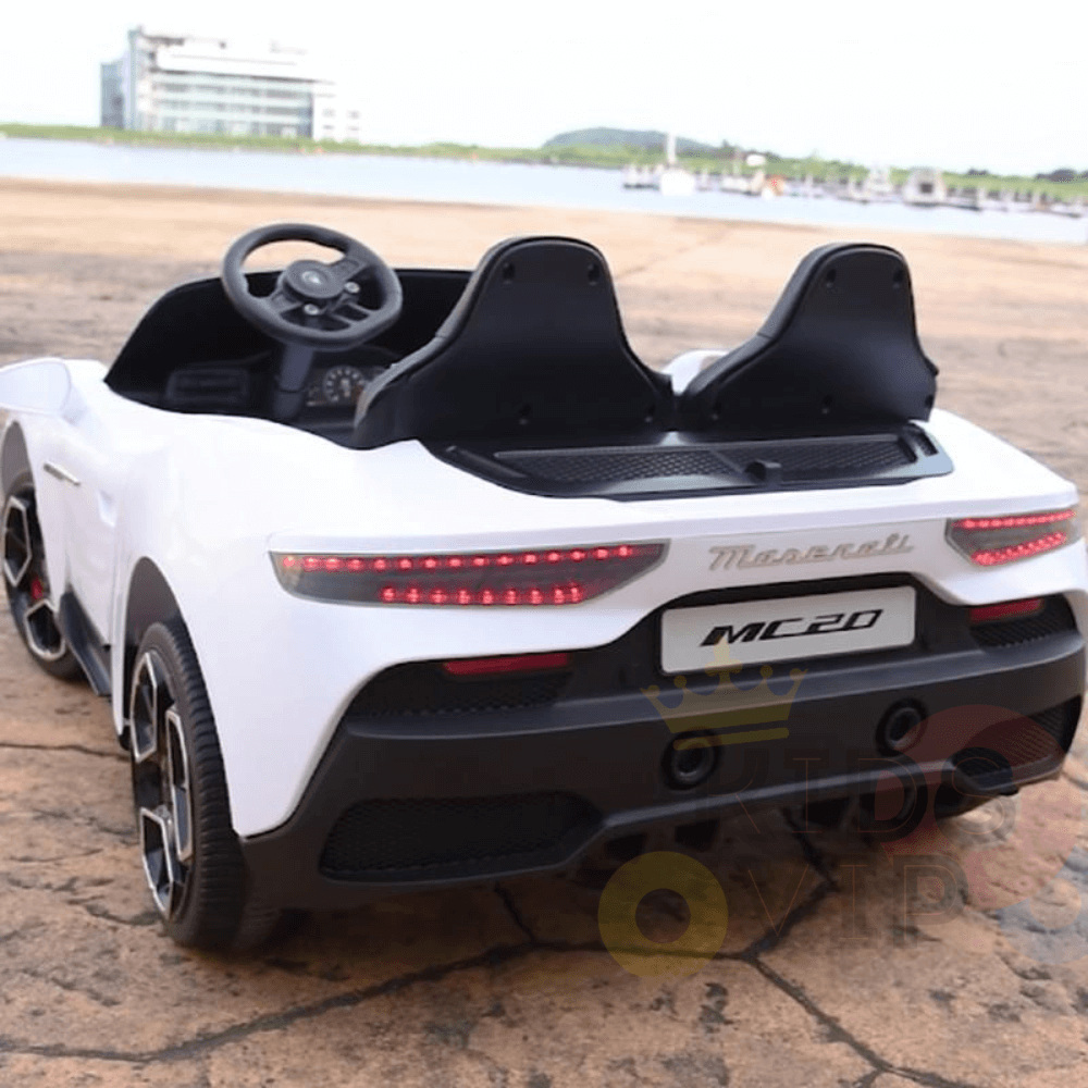 A white XXL 24V/105W Maserati MC20 toy car, featuring premium leather seats and rubber tires, is parked on a stone surface with water and a modern building in the background.