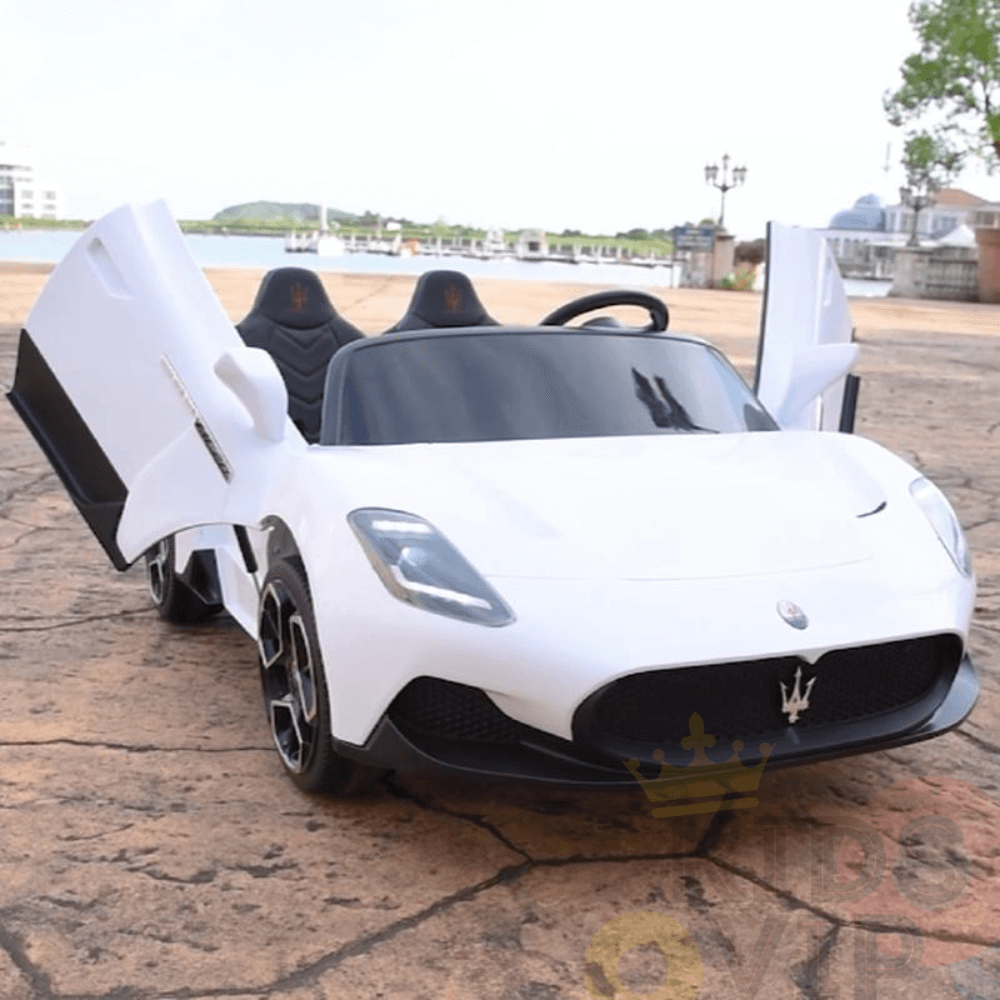 A white XXL 24V Maserati MC20 toy car with butterfly doors open sits on a brick surface, featuring black leather seats and the iconic grille. With trees and buildings as a backdrop, this premium 2-seater ride-on is luxury epitomized.