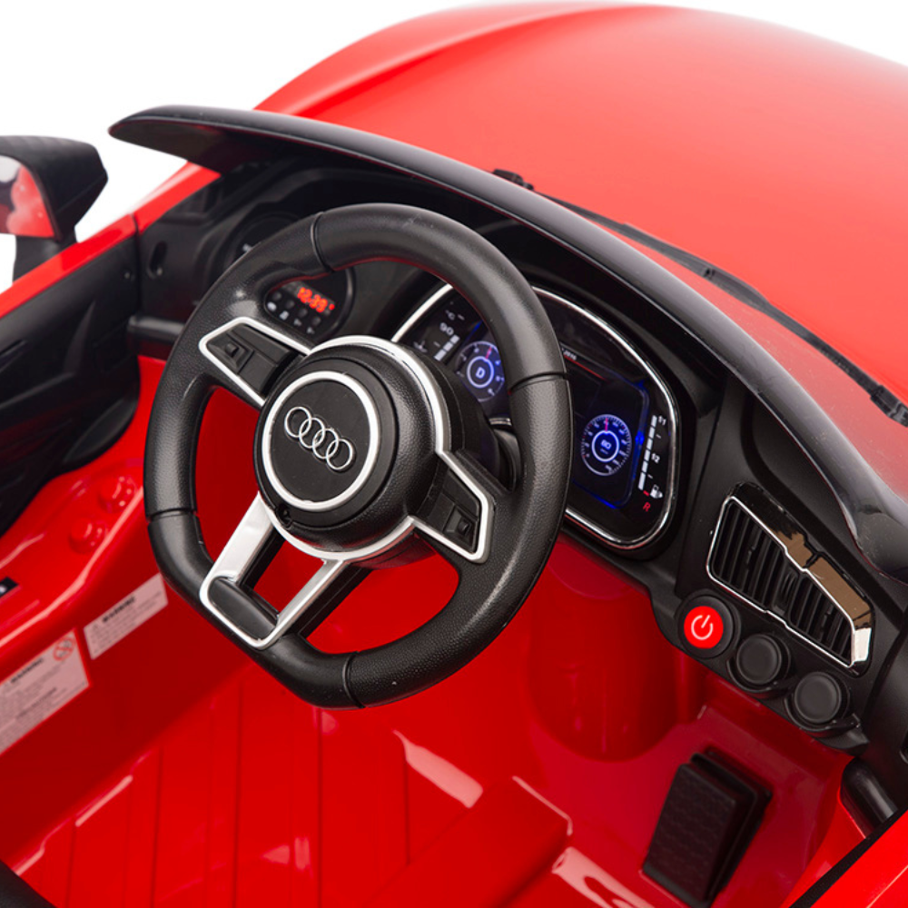 Close-up of a child-sized Red 12V Audi R8 Licensed 1-Seater Ride-On Cars interior, features a black steering wheel with an Audi logo, digital dashboard with illuminated displays, control buttons, and upgraded EVA rubber wheels powered by a 12V battery.