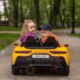 Two children ride in a yellow XXL 24V /105W Maserati MC20 toy car with leather seats and rubber tires. The girl wears a purple jacket; the boy sports an orange jacket and blue cap. Trees and a lamppost enhance the scene as they cruise their premium playground edition car.